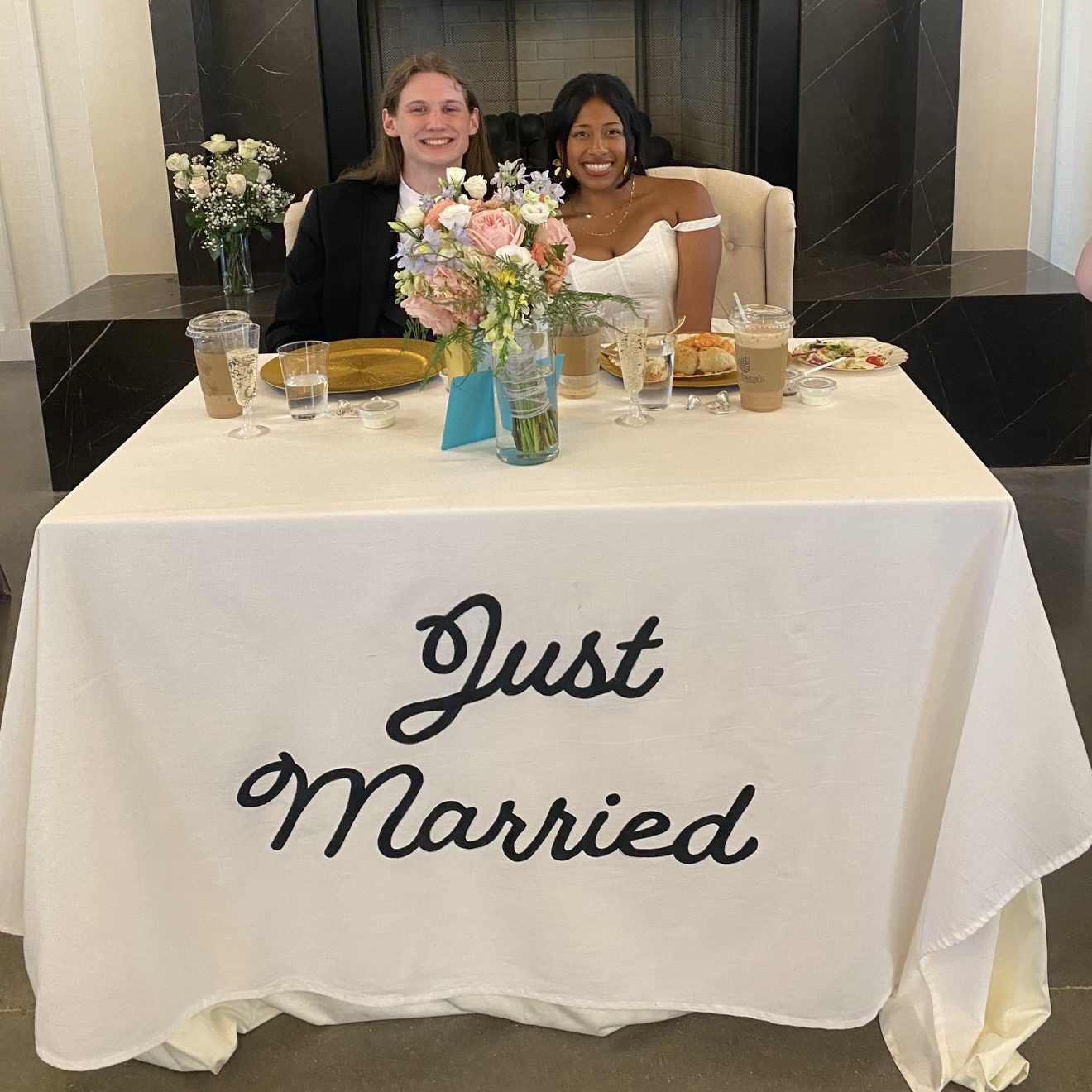 Newlyweds smiling behind a "Just Married" table with flowers and drinks.